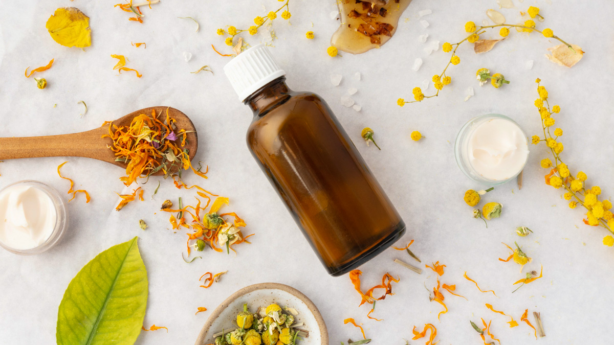 Braune Glasflasche mit weißem Verschluss, umgeben von getrockneten Blüten, Creme in kleinen Gläsern, Honig und einem grünen Blatt auf weißem Untergrund